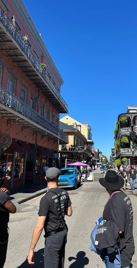 Friends in black trip shirts and a wide-brimmed hat walk down a sunny French Quarter street lined with iron balconies.