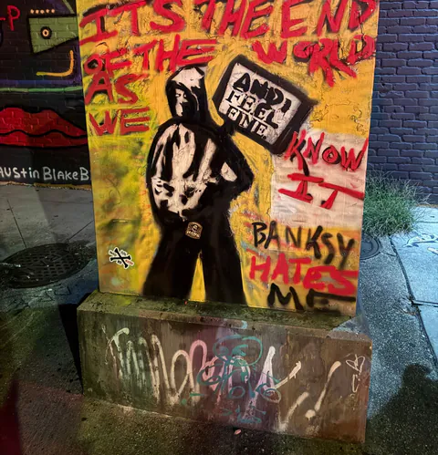A yellow stencil-art utility box reading Banksy Hates Me, with a small Haws sticker on the painted figure's belt.