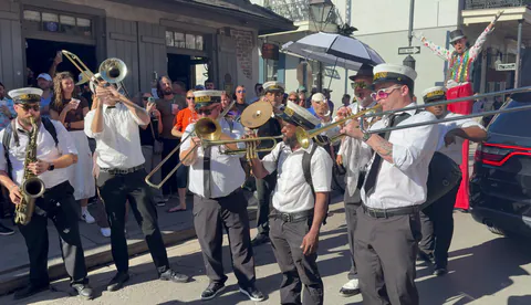 A brass band in white shirts and Big Fun caps blasts trombones, trumpets and a saxophone in the street as a stilt walker in a sequined vest throws both arms up behind a white umbrella and a crowd watches.