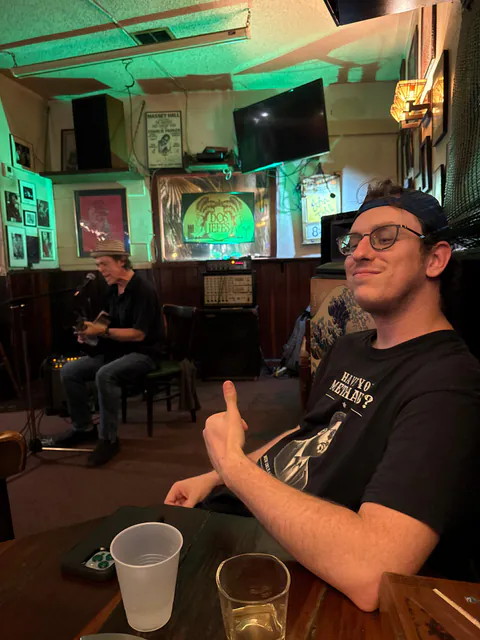 A man in a Have You Met Haws? shirt gives a thumbs-up while a guitarist sings and plays in a wood-paneled bar.