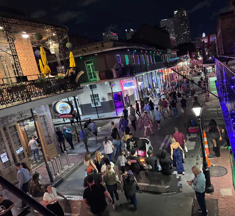 A night view from a balcony over a crowded Bourbon Street lined with iron galleries, neon signs and yellow umbrellas.