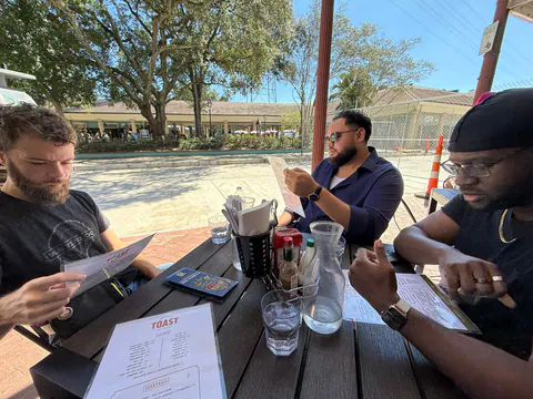 Three men study menus at a sunny outdoor brunch table under big oak trees.