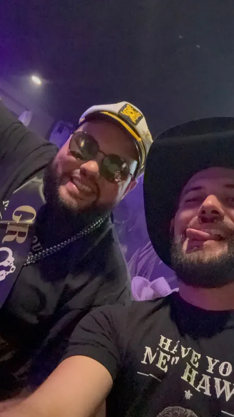 Night selfie of the groom in a captain's hat, sunglasses and GROOM sash beside a friend in a cowboy hat and Haws shirt.