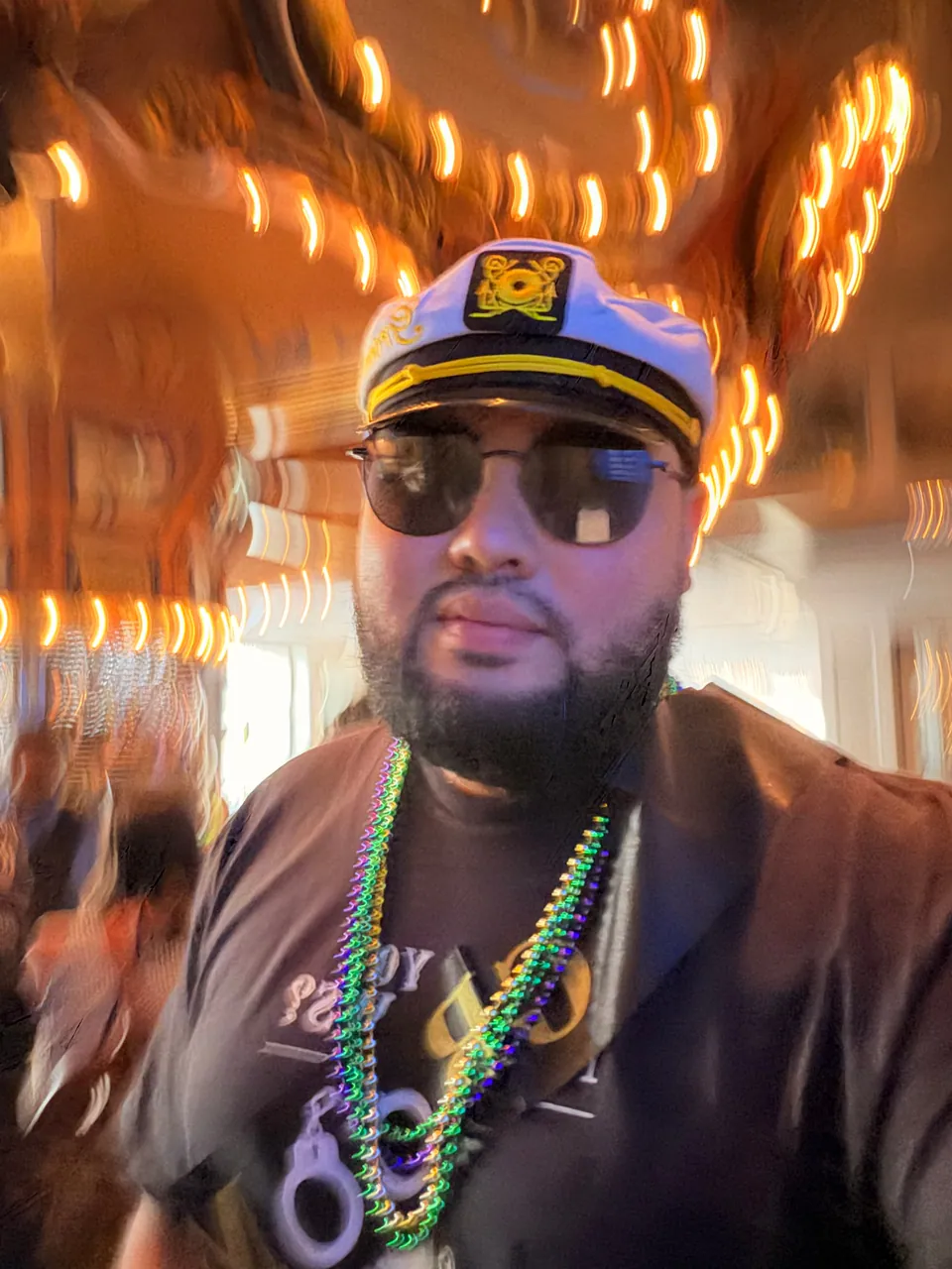 The groom in a white captain's hat, sunglasses and Mardi Gras beads takes a selfie under streaking golden carousel lights.