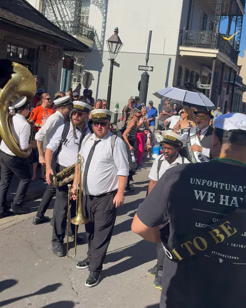 Seen from behind, the groom in a captain's hat, an Unfortunately, We Have shirt and a black-and-gold sash faces a brass band in white shirts and Big Fun caps on a sunny street corner.