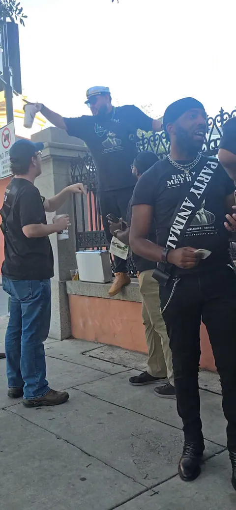 The groom in a captain's hat stands on a ledge by an iron fence raising a cup as friends in Haws shirts, one in a Party Animal sash, mingle below.