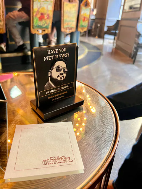 A Have You Met Haws? table card on a round glass bar table beside a Carousel Bar napkin, bar seats blurred behind.