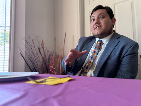 A man in a blue plaid suit and bright checkered tie gestures mid-story at a table with a purple tablecloth.