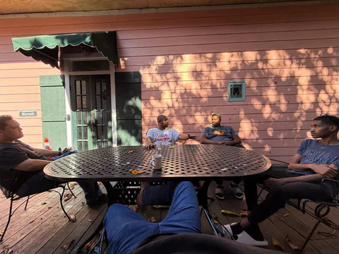 Friends talk around a metal patio table on a wooden deck as dappled morning sun hits a pink clapboard wall.