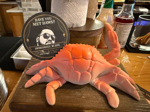 A Have You Met Haws? coaster propped behind an orange crab figurine on a wooden bar.