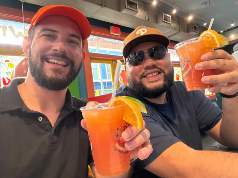 Two smiling friends in caps raise orange iced drinks with orange-slice garnishes in a brightly painted restaurant.