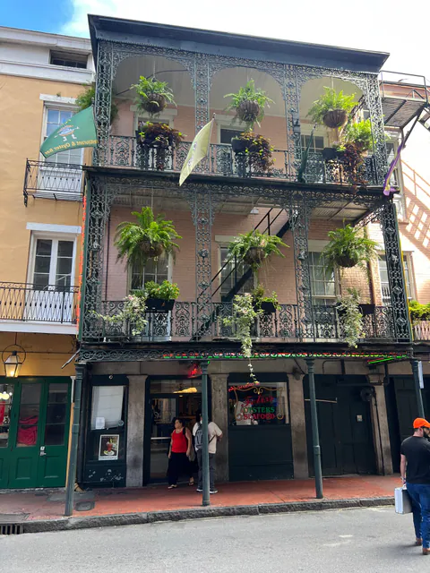 A pink French Quarter building with fern-hung iron balconies above an oyster bar as a man with a silver briefcase walks by.