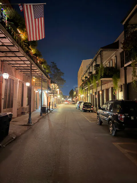 A quiet French Quarter street at night, lined with fern-draped iron balconies, glowing lamps and an American flag.