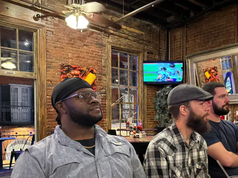 Three men watch a game in a brick-walled bar with tall wood-framed windows, a football broadcast on the TV behind them.