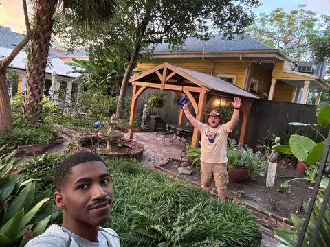 A friend takes a deadpan selfie while a man in a wide-brimmed hat cheers with both arms up in a lush courtyard garden.