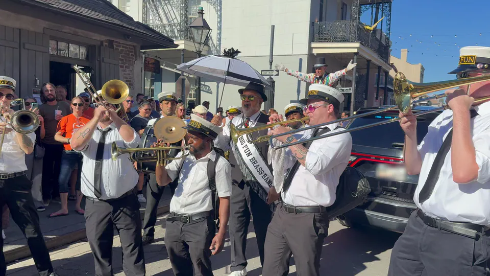 A grand marshal in a black hat and Big Fun Brass Band sash holds up a white umbrella among trumpet and trombone players while a stilt walker spreads both arms above the crowd.