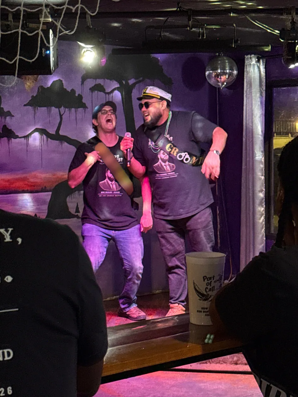 The groom, in a captain's hat and GROOM sash, belts out karaoke with a friend in a cap and glasses on a purple-lit stage.
