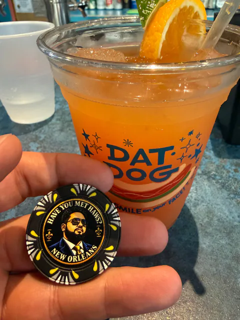 A hand holds a black Have You Met Haws? New Orleans poker chip in front of an orange frozen drink in a Dat Dog cup.