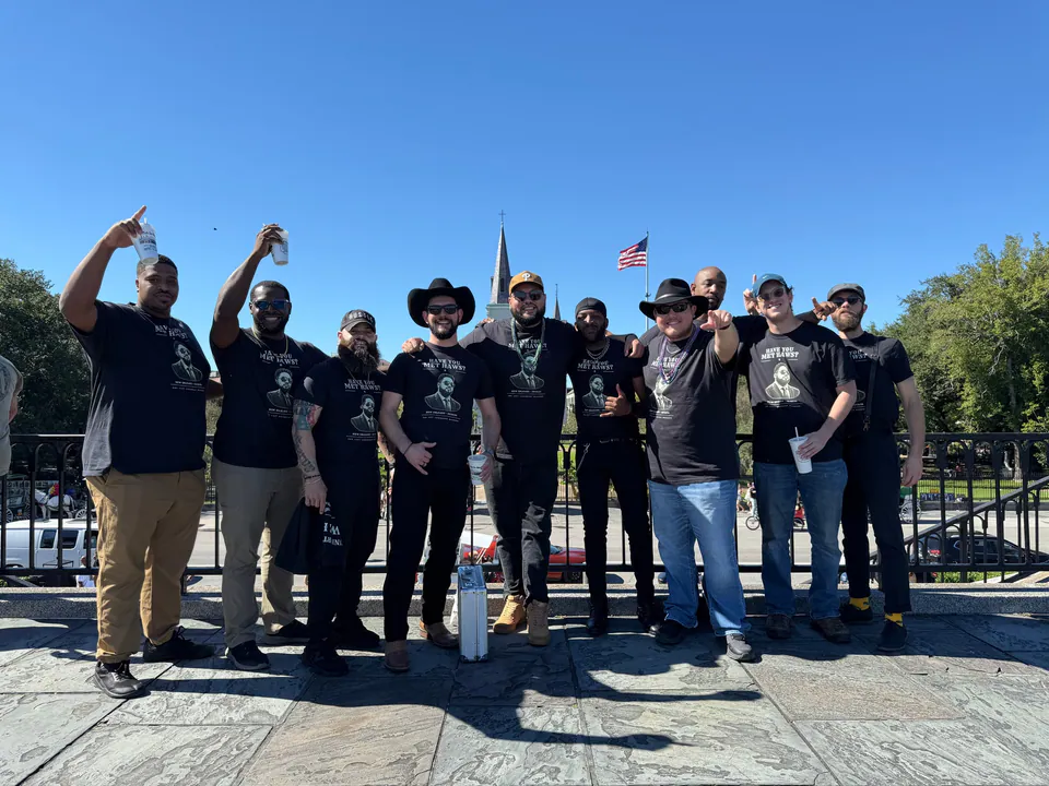 Ten friends in matching black Have You Met Haws? shirts cheer and raise cups before St. Louis Cathedral under a blue sky.
