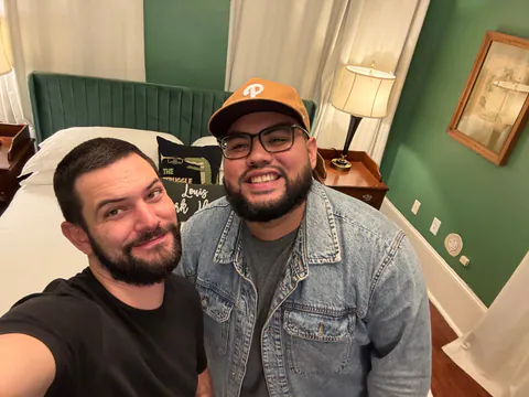Two bearded friends, one in a denim jacket and cap, smile for a late-night selfie in a green room with a trumpet pillow.