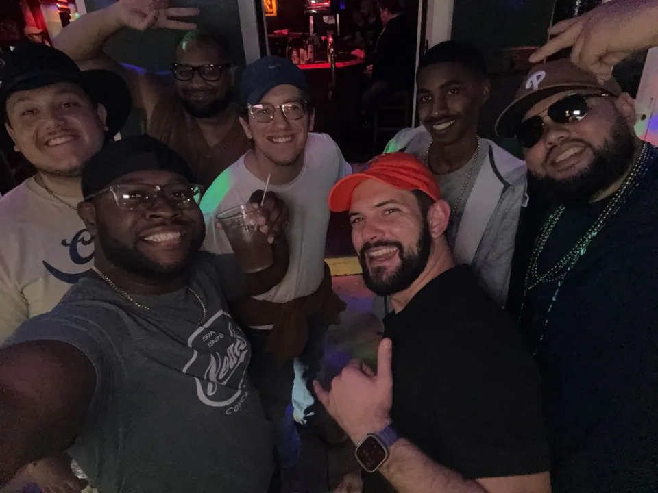 Seven grinning friends in caps and beads squeeze into a late-night bar selfie, throwing shakas and peace signs.