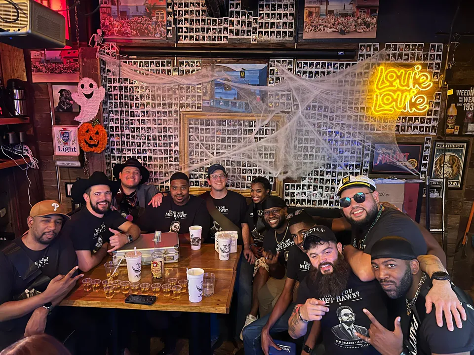 Eleven friends in matching Have You Met Haws? shirts crowd a bar table under a Louie Louie neon sign, the groom in a captain's hat at right.