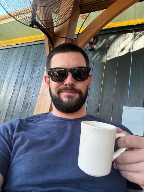 A smiling bearded man in sunglasses raises a white coffee mug under a wooden patio roof.