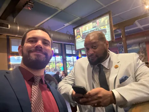 Two men in suits and ties, one in a striped seersucker jacket, pose for a selfie in a bar with a TV behind them.