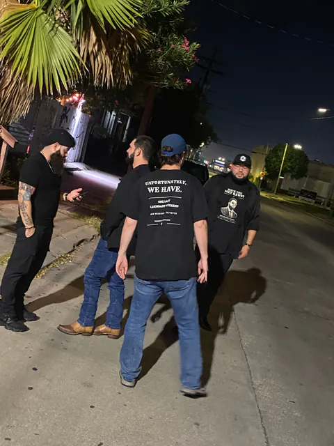 Four friends talk on a night sidewalk under a palm tree; one shirt back reads Unfortunately, We Have., another shirt front reads Have You Met Haws?