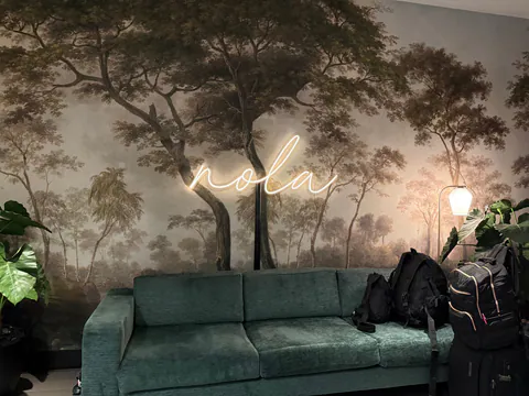 A glowing neon nola sign on a forest mural above a green sofa piled with backpacks.