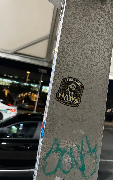 An I Bought Haws a Drink sticker showing a man in sunglasses and a suit, stuck on a metal pole at night.