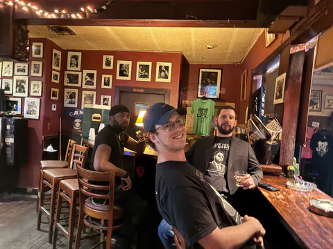 Three friends at a photo-lined bar, one in a blazer over a Have You Met Haws shirt.