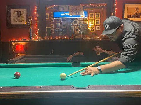 A man in a black cap lines up a pool shot on a green table while a friend sips a drink on the couch behind.