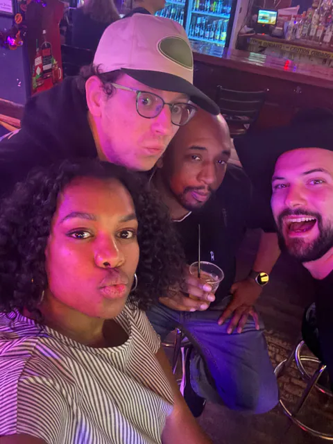 Four friends squeeze into a purple-lit bar selfie, two pulling duck faces and one laughing.
