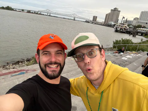 Two friends take a selfie on the Mississippi riverfront steps with a steel bridge and riverboat behind them.