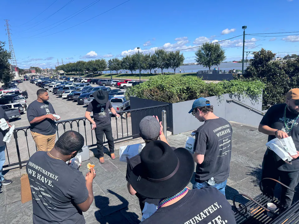 Friends in matching black shirts reading Unfortunately, We Have share beignets on a sunny riverfront terrace.