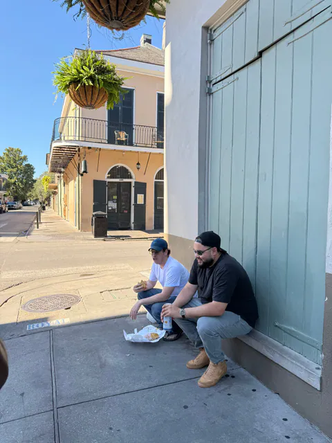 Two friends eat lunch on a sunny Royal Street sidewalk, sitting against green shutters beneath a hanging fern.