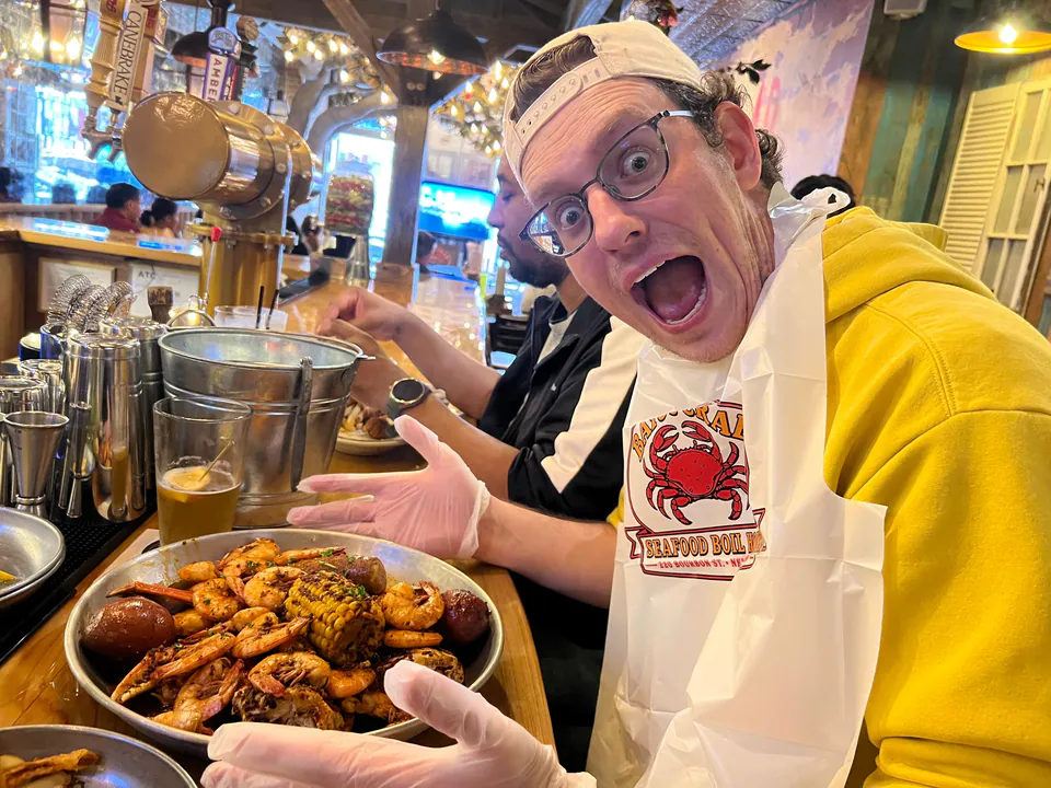 A man in a crab bib and plastic gloves makes a wide-mouthed face over a tray of boiled shrimp, corn and potatoes.