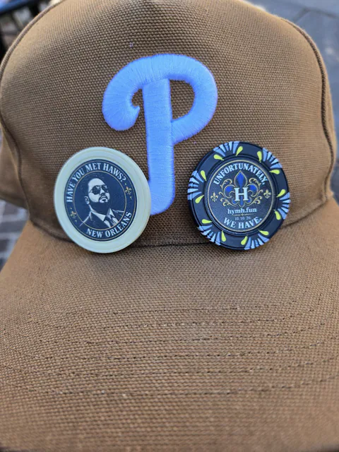 Two Haws souvenir chips, one with a suited portrait and one with a fleur-de-lis and hymh.fun, rest on the front of a brown ball cap.