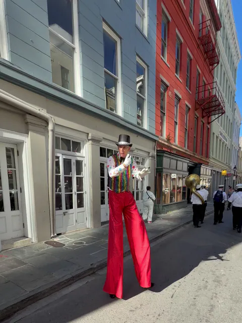 A stilt walker in a top hat, sequined vest and red trousers gestures toward the camera on a sunny French Quarter street as a brass band with a sousaphone marches behind.