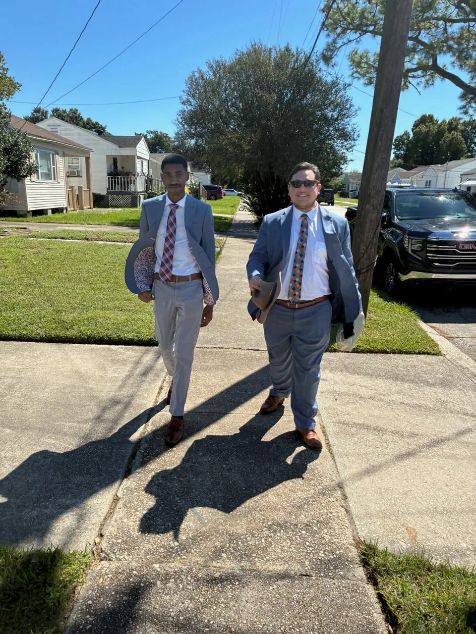 Two men in light blue suits and patterned ties stroll down a sunny neighborhood sidewalk, hats in hand.