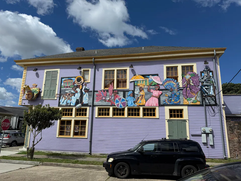 A lavender two-story building covered in a mural of brass band players, parasols and feathered Mardi Gras Indian suits.