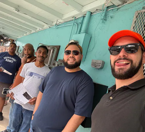 Five friends in T-shirts and caps squeeze into a selfie against a turquoise restaurant wall, menus in hand.