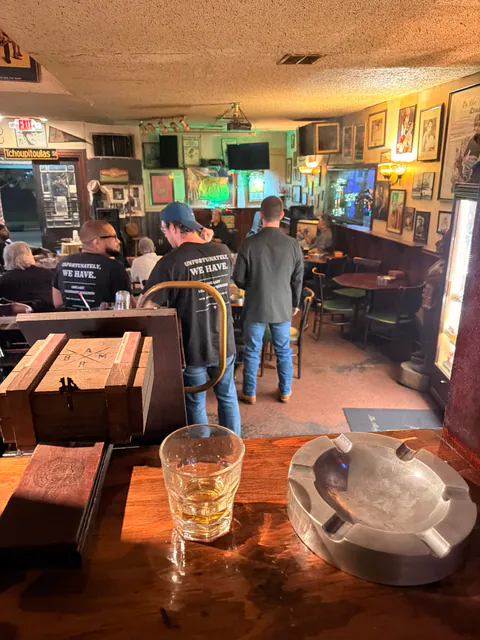 Friends in shirts reading Unfortunately, We Have gather in a photo-lined dive bar, seen past a glass and an ashtray.